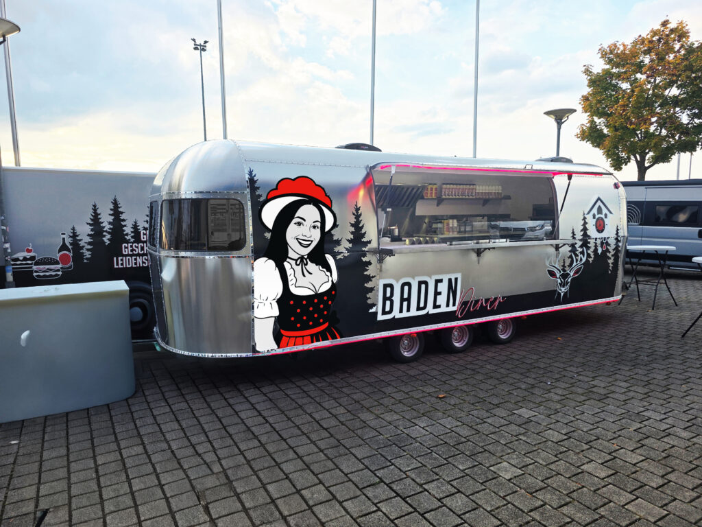 Silberner Retro-Verkaufswagen mit der Aufschrift „BADEN Diner“. Auf der Seite ist eine Grafik einer lächelnden Frau in Tracht zu sehen. Der Wagen steht auf einem gepflasterten Platz, die Verkaufsklappe ist geöffnet und gibt den Blick auf die Küche frei.