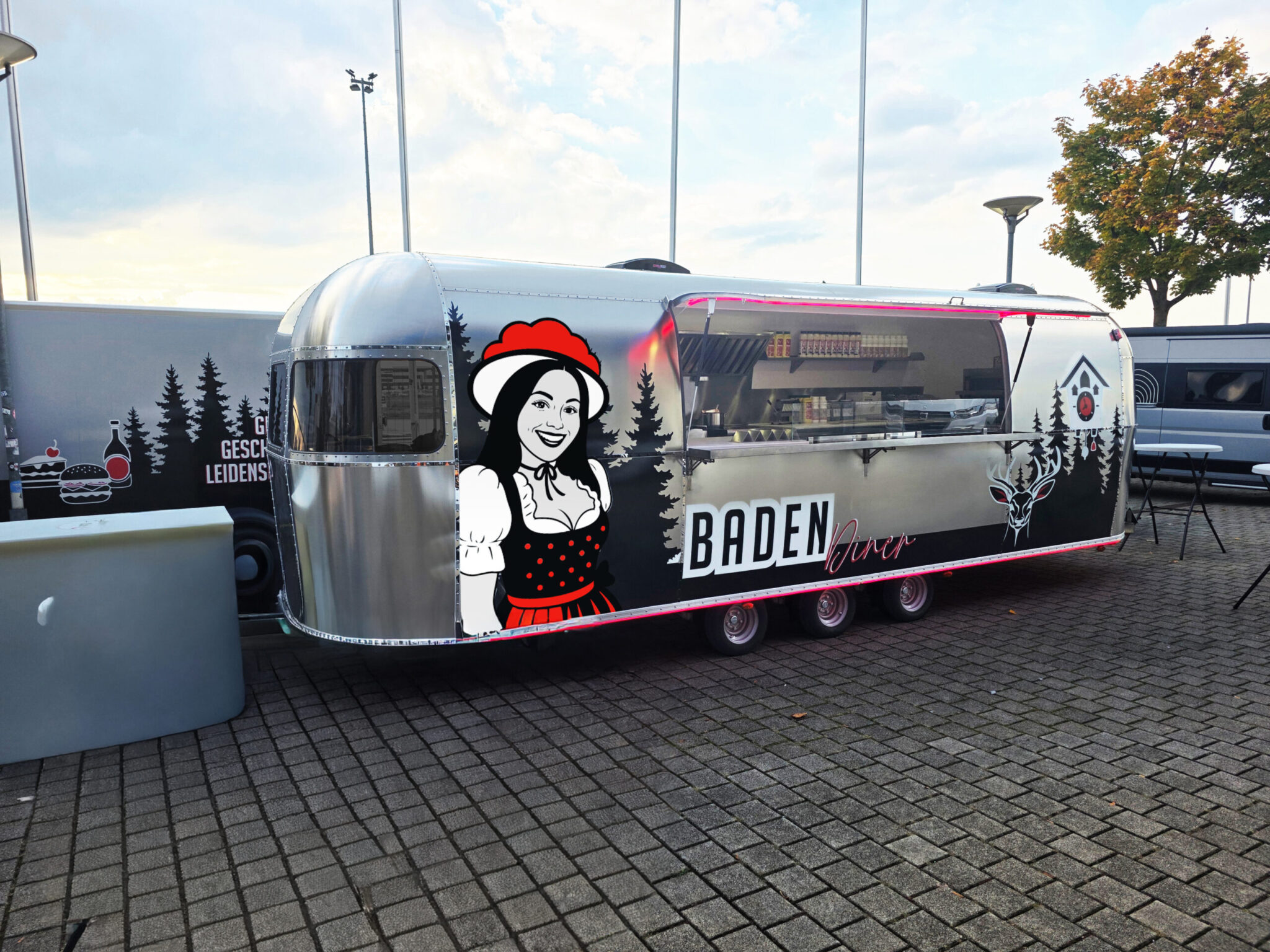 Silberner Retro-Verkaufswagen mit der Aufschrift „BADEN Diner“. Auf der Seite ist eine Grafik einer lächelnden Frau in Tracht zu sehen. Der Wagen steht auf einem gepflasterten Platz, die Verkaufsklappe ist geöffnet und gibt den Blick auf die Küche frei.